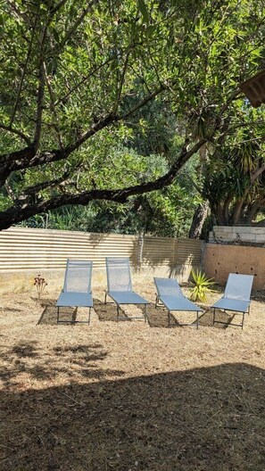 A shaded outdoor area with metal lounge chairs, a concrete wall, and dense foliage.