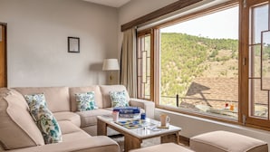 A living room with a beige sofa, a coffee table, and a view of a hilly landscape through large windows. - The Friendship Lane (Salt)