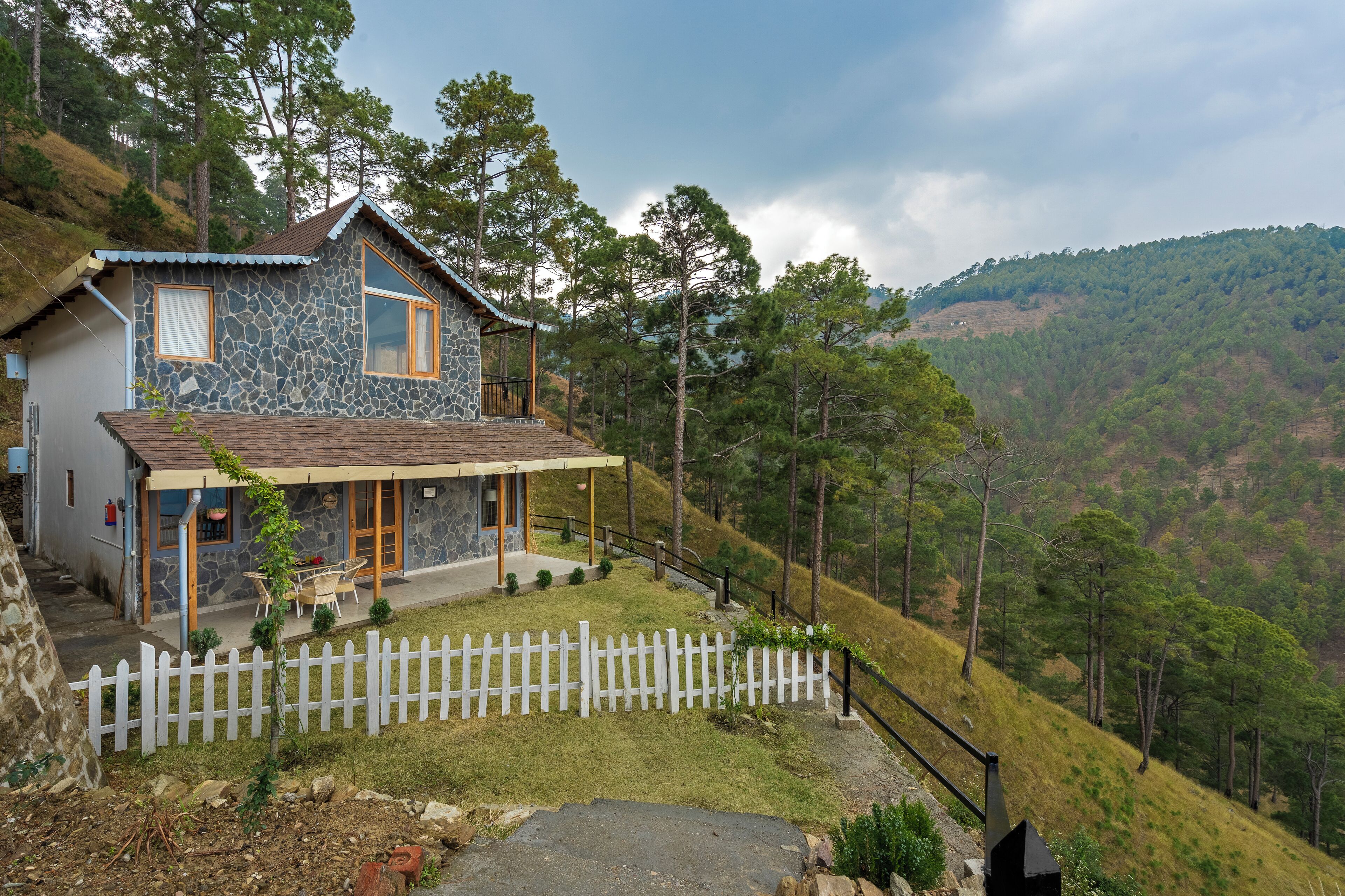 A stone house with a porch, surrounded by a white picket fence and a view of a hilly landscape.