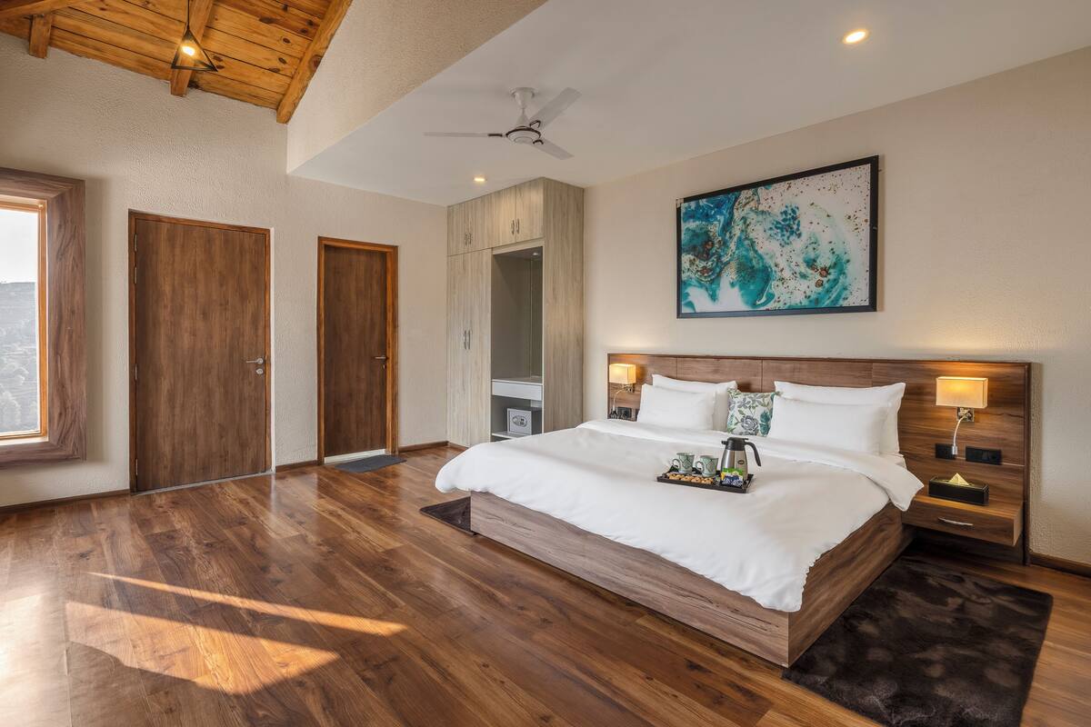 A spacious bedroom with a large bed, wooden flooring, a ceiling fan, and a painting on the wall.