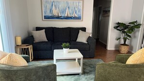 Una sala de estar con un sofá gris oscuro, una mesa de centro blanca y dos sillones verdes. Un cuadro de veleros cuelga en la pared, y hay una planta en maceta en el suelo.