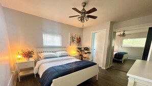 Un dormitorio con cama, lámparas de noche, ventilador de techo, ventana con persianas y puerta que da a otra habitación.