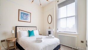 Une chambre bien rangée, avec un grand lit, une table de chevet avec une lampe, un tableau encadré au mur et une fenêtre laissant entrer la lumière naturelle.