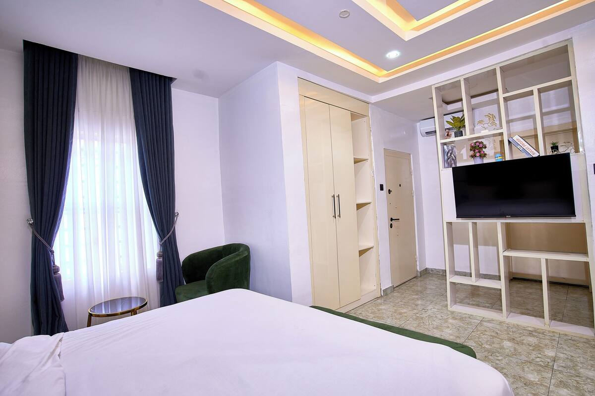 Une chambre d’hôtel avec un lit, un fauteuil vert, une armoire, un téléviseur posé sur un meuble et une petite table.