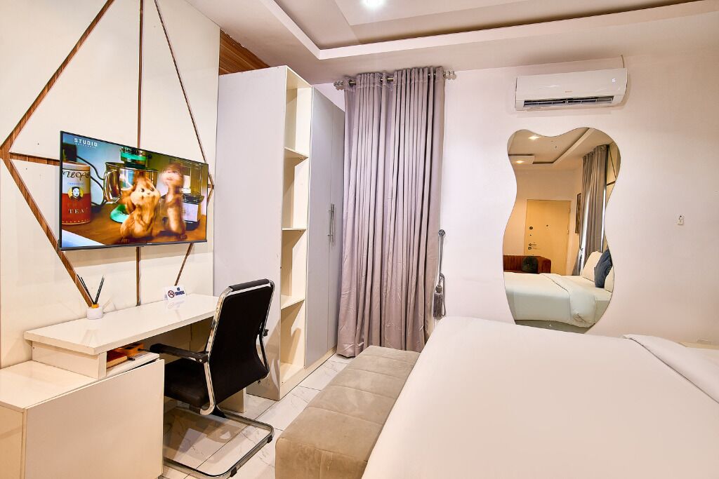 A modern hotel room with a bed, a desk with a chair, a television, and a bathroom visible through an archway.