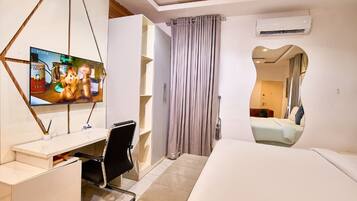 A modern hotel room with a bed, a desk with a chair, a television, and a bathroom visible through an archway.