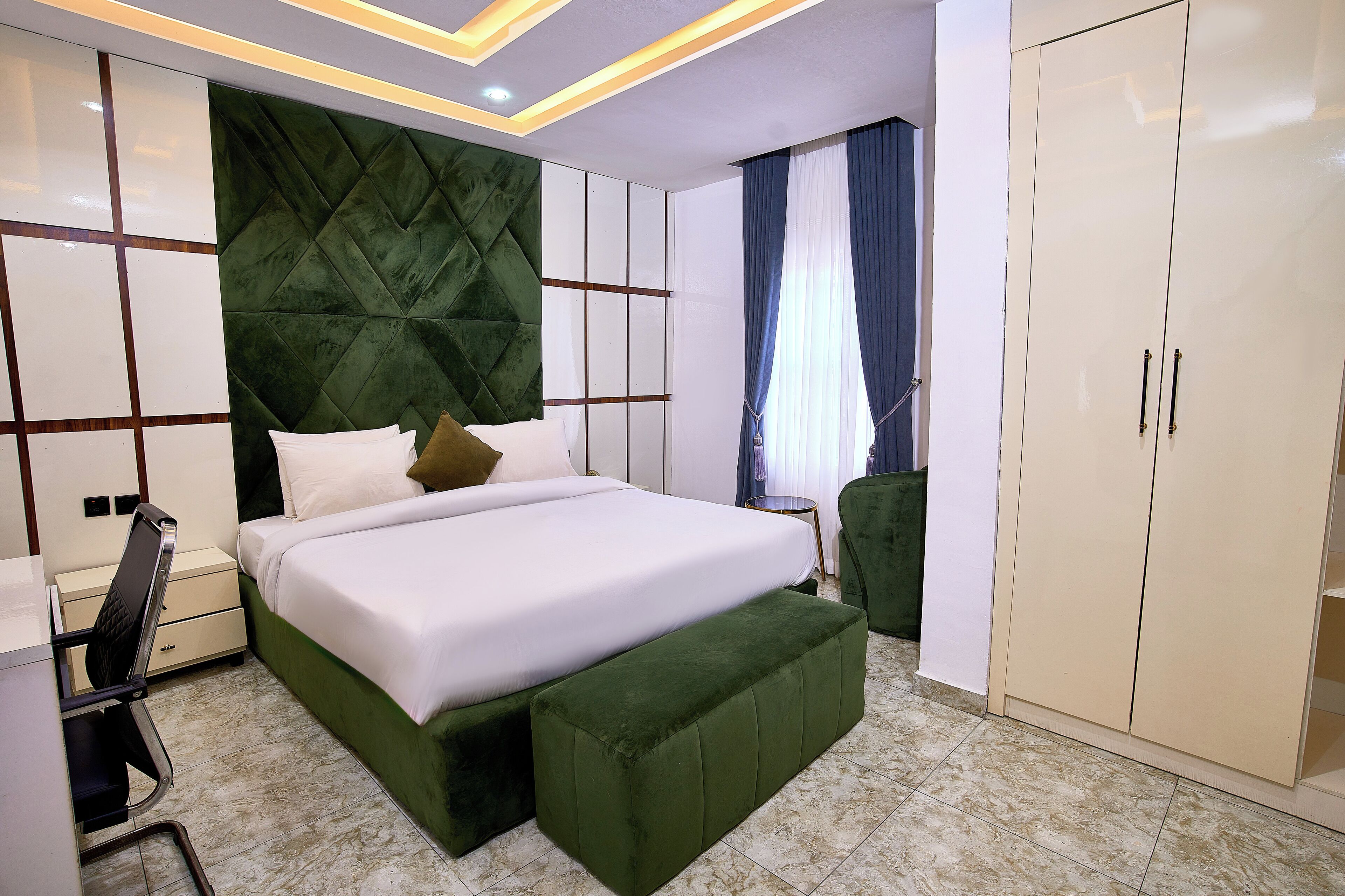 Une chambre à coucher comprenant un lit, un banc recouvert d’un tissu vert, une armoire blanche et un bureau avec une chaise.