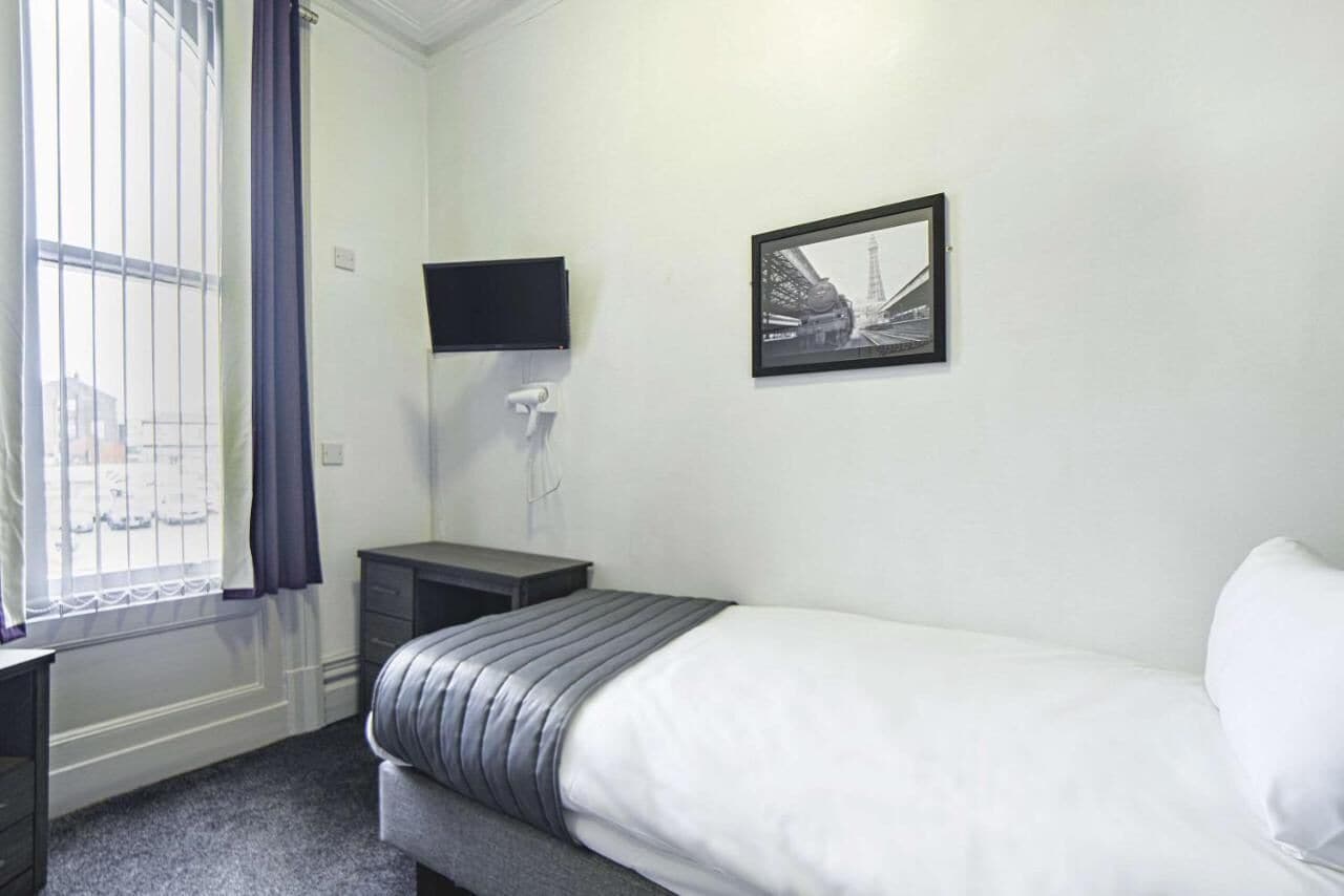 Ein Schlafzimmer mit einem Bett, einem Schreibtisch, einem Stuhl, einem Fenster mit Vorhängen, einem Flachbildfernseher und einem gerahmten Bild an der Wand.