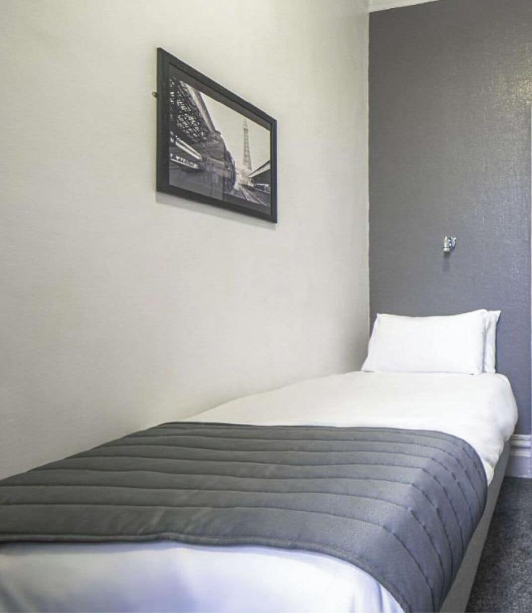 Uma cama de solteiro com cabeceira cinza e roupa de cama branca em um quarto com parede de destaque cinza e um quadro de uma ponte.
