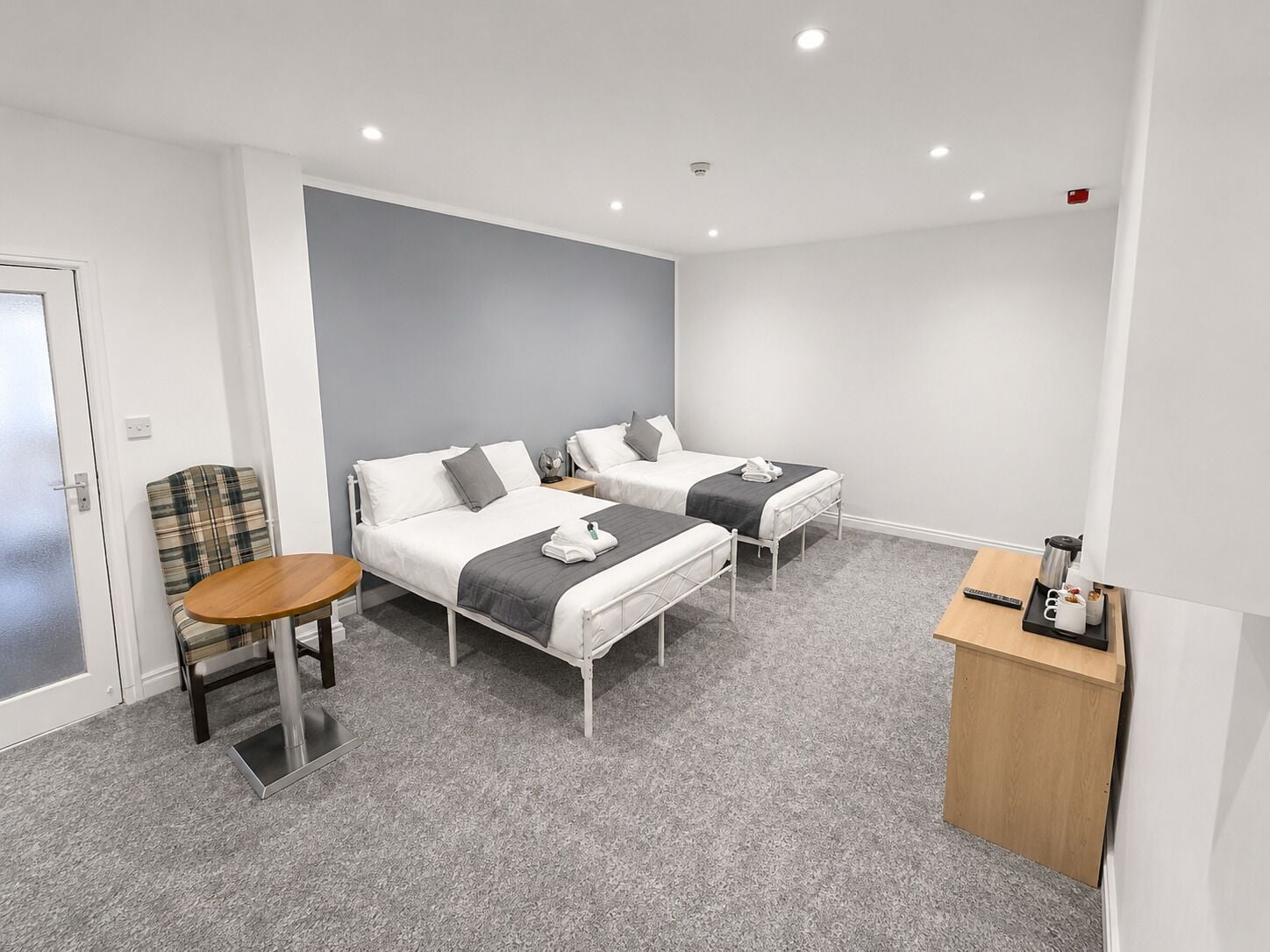 A hotel room with two beds, a small table, a chair, and a kitchenette area.