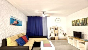 Una sala de estar con un sofá, una mesa de centro, un televisor, un ventilador de techo, un reloj y una estantería.