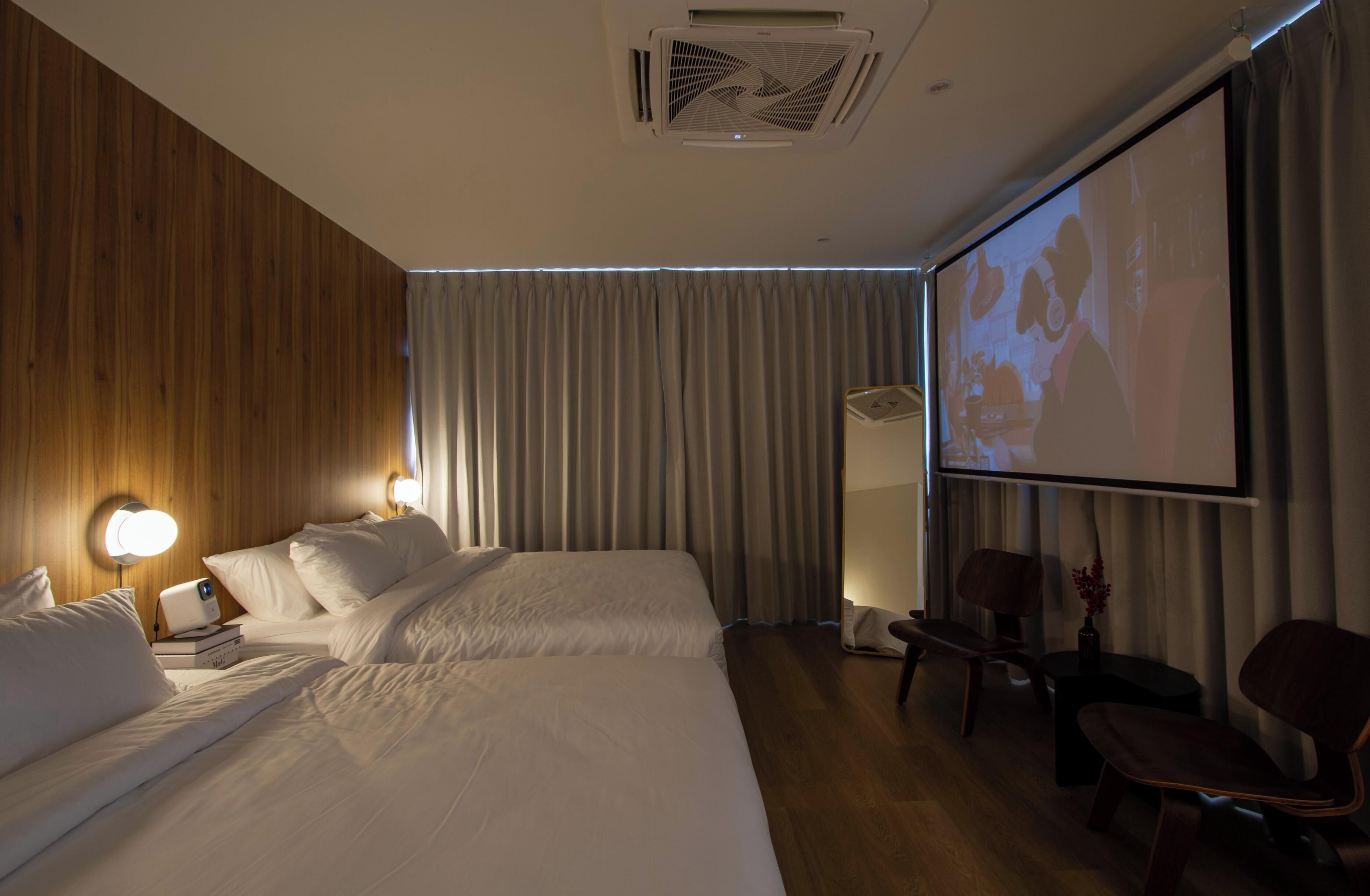 A hotel room with two beds, a projector screen, wooden panel walls, and a seating area with chairs.