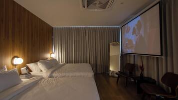 A hotel room with two beds, a projector screen, wooden panel walls, and a seating area with chairs.