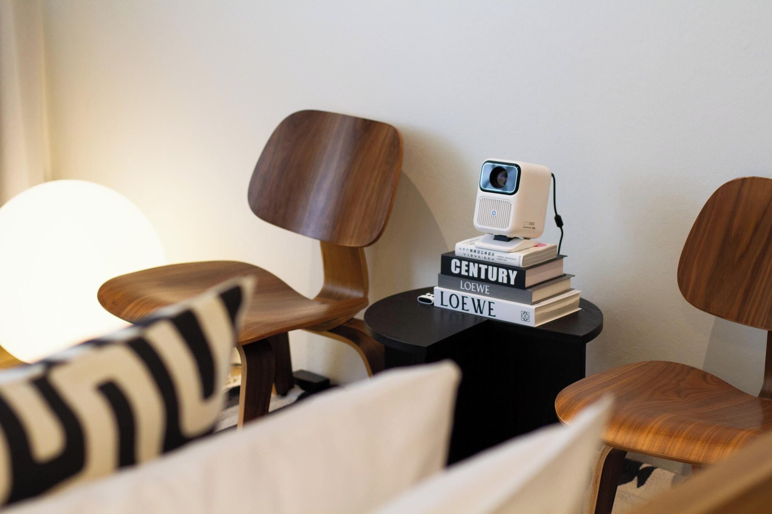 Una habitación moderna con sillas de madera, una pequeña mesa negra con una cámara web y libros, y una lámpara grande.