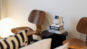 Una habitación moderna con sillas de madera, una pequeña mesa negra con una cámara web y libros, y una lámpara grande.