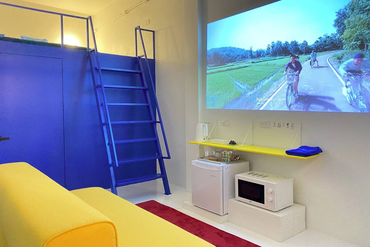 Une chambre équipée d’un lit superposé, d’une échelle, d’un four à micro-ondes, d’un petit réfrigérateur et d’un téléviseur diffusant une scène de course cycliste.