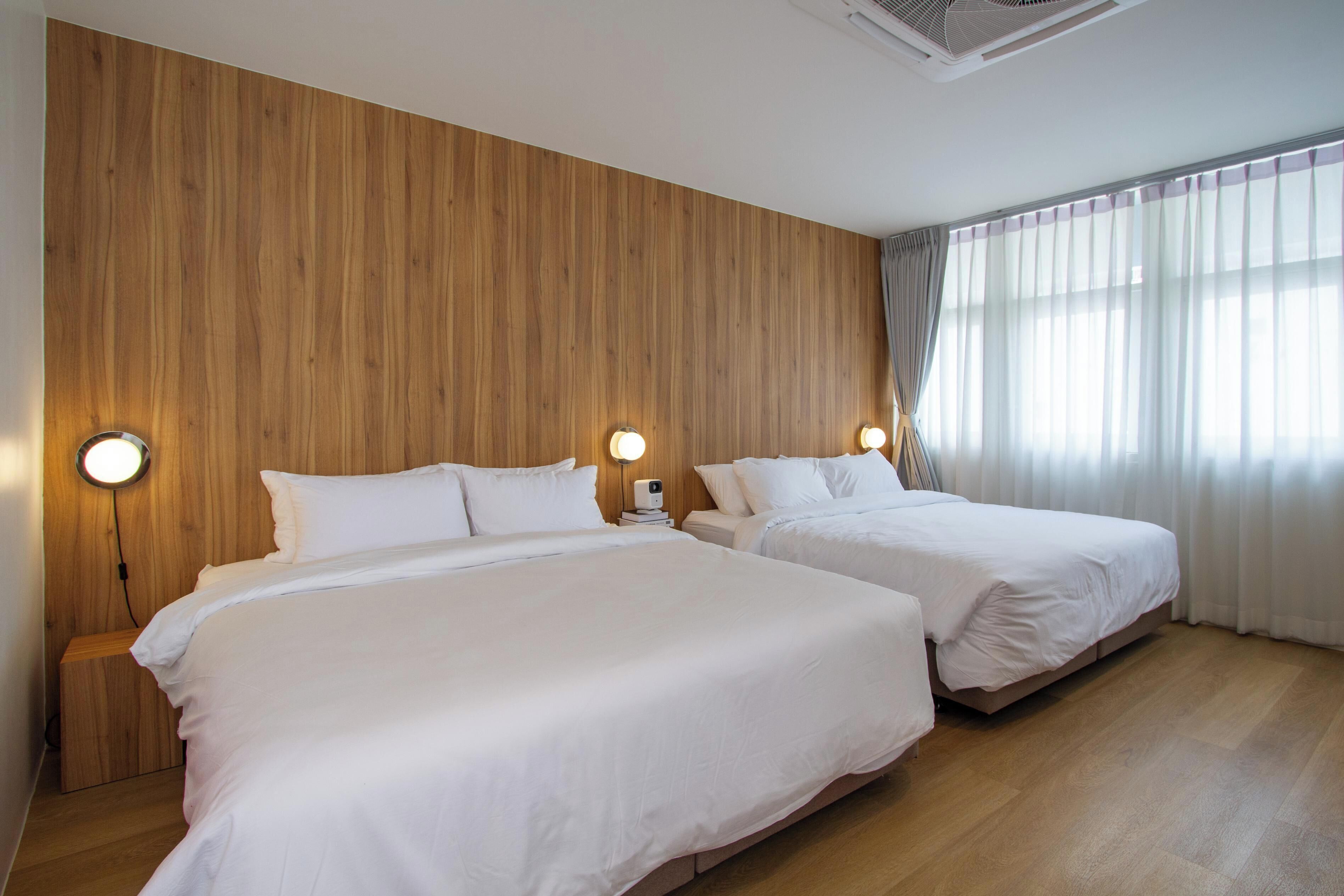 A hotel room with two beds, wooden panel walls, and a large window with sheer curtains.