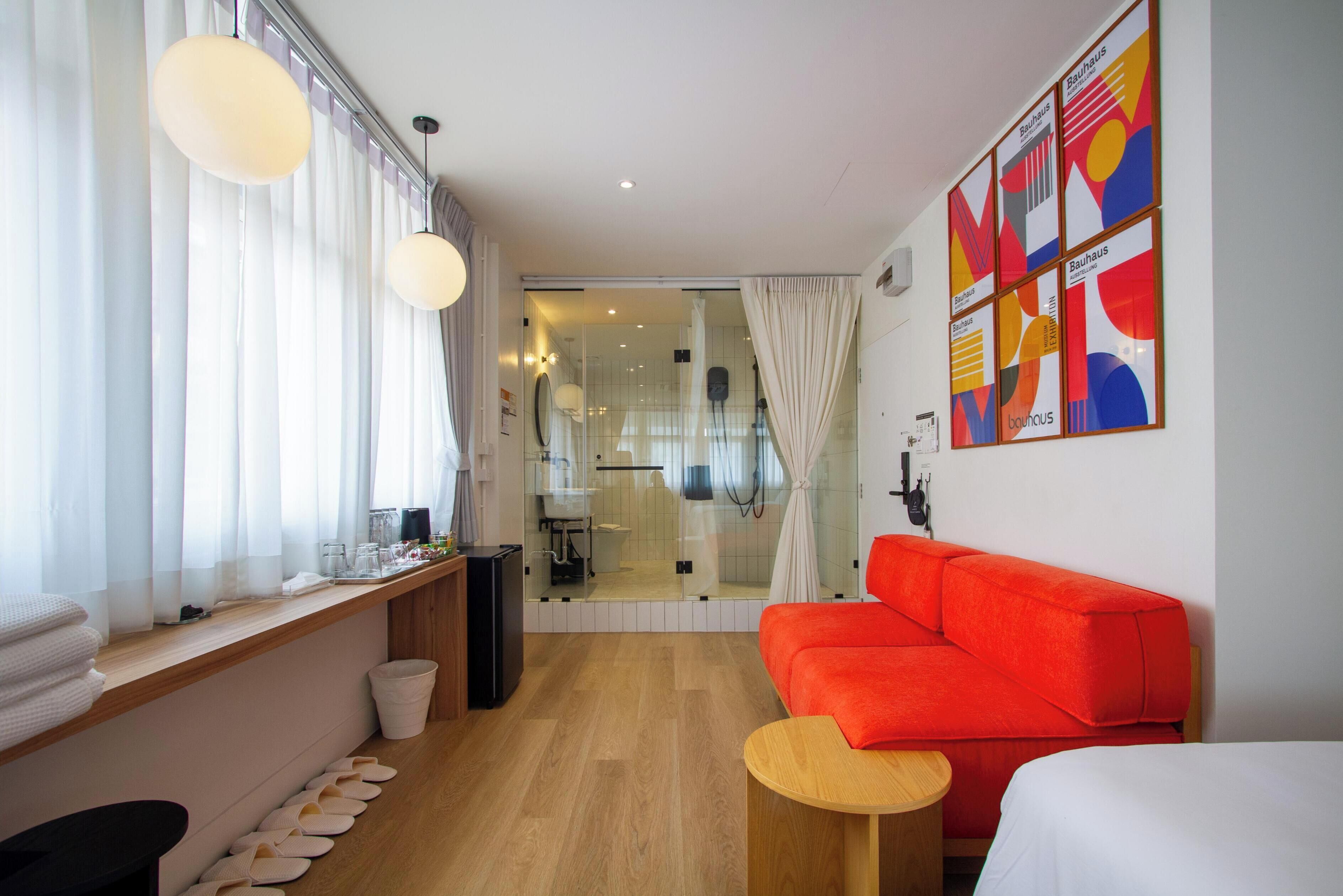 Una habitación de hotel moderna con suelo de madera, un sofá rojo, una mesita redonda y un ventanal con cortinas translúcidas.