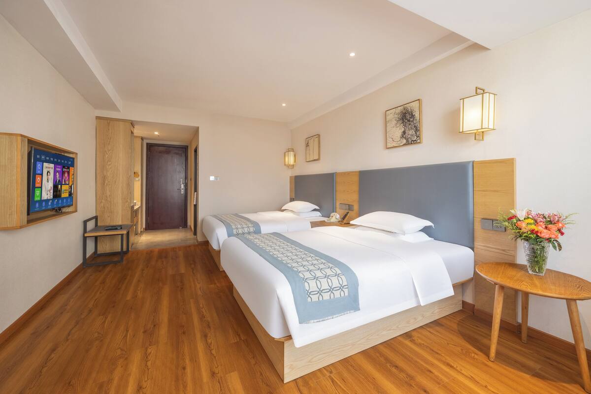 Une chambre d’hôtel avec un plancher en bois, deux lits avec une literie bleue et blanche, une petite table sur laquelle est posé un vase de fleurs et un téléviseur fixé au mur.