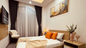 A modern hotel room with a large bed, a wooden headboard, a fluffy white rug, a bedside table with a lamp, and a wall-mounted TV. - căn hộ biển (Thành phố Phú Quốc)