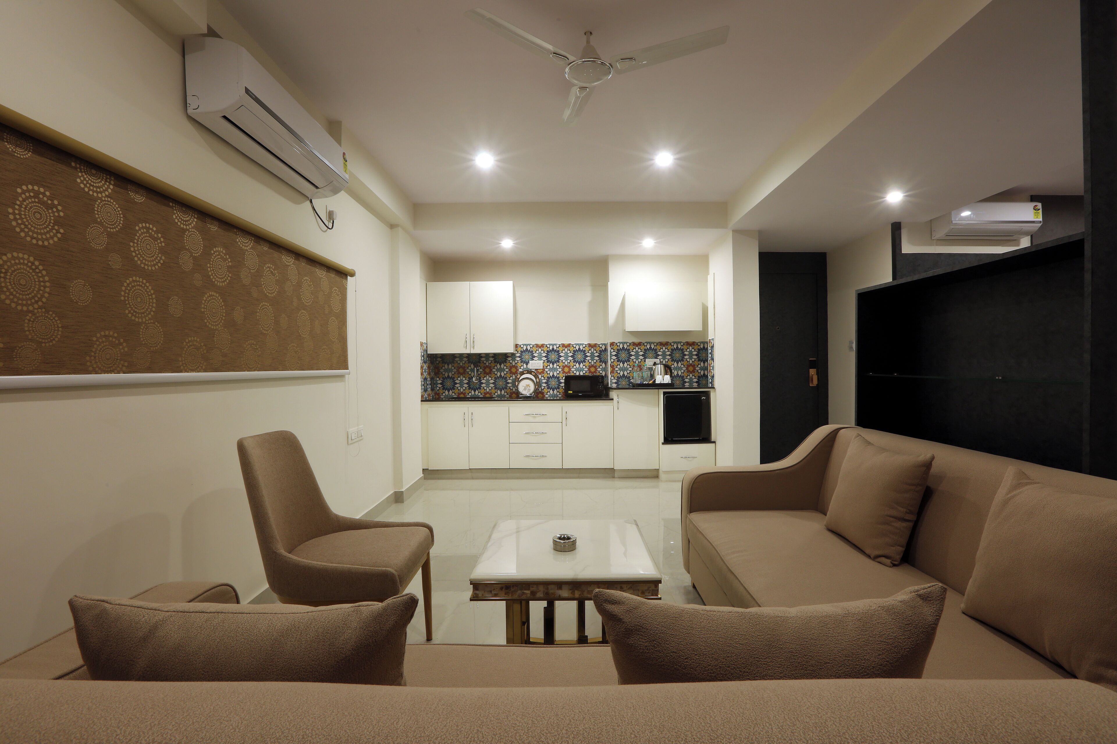 A modern living room with a sofa, a coffee table, and a TV. There is a kitchen area with cabinets and a microwave.