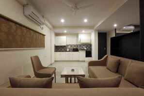 A modern living room with a sofa, a coffee table, and a TV. There is a kitchen area with cabinets and a microwave. - The Hoften Sterling Plaza Near Kempegowda International Airport (Bengaluru)