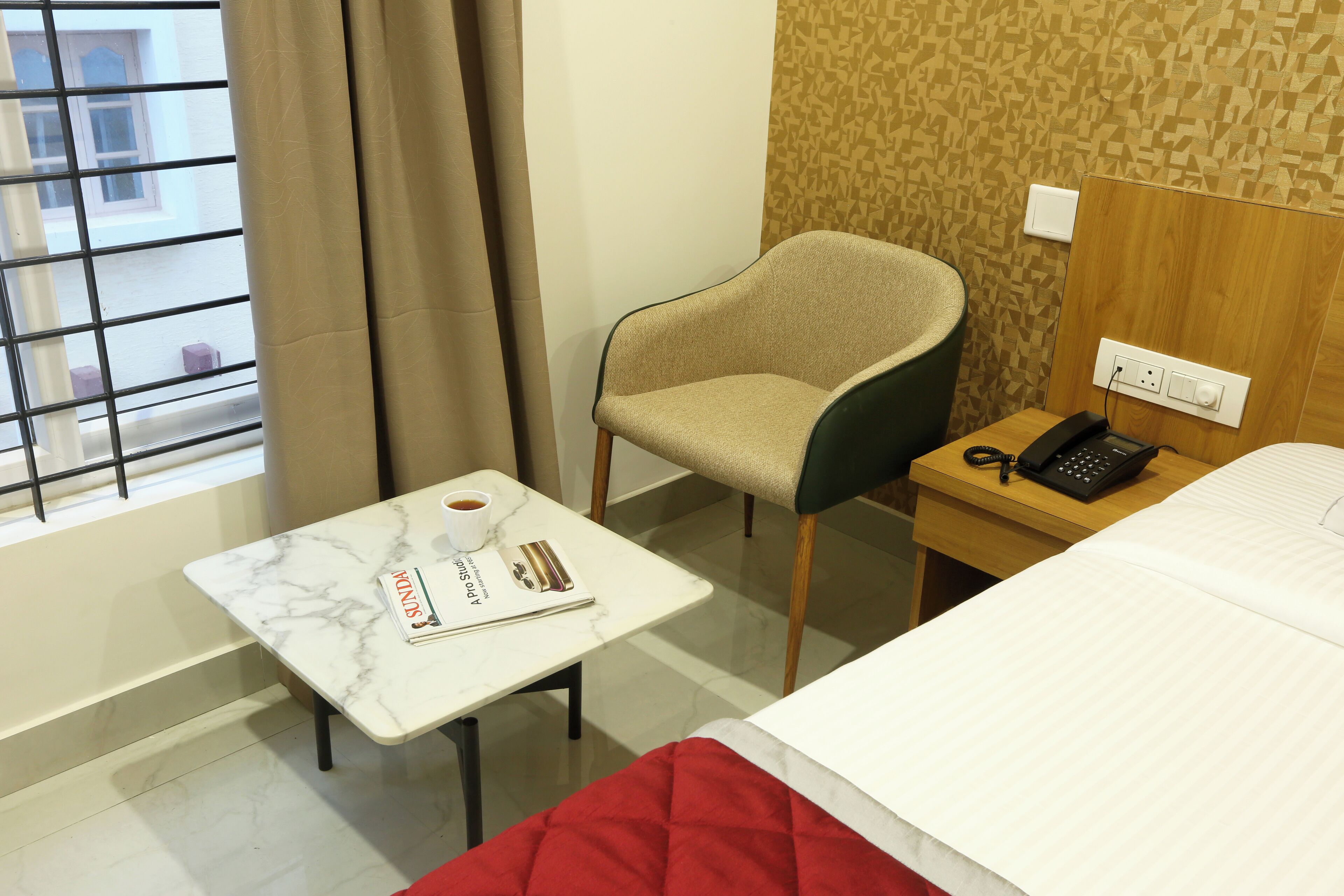 A hotel room with a bed, a small table with a magazine, a chair, a telephone, and a window with curtains.
