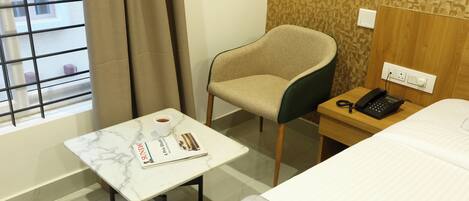 A hotel room with a bed, a small table with a magazine, a chair, a telephone, and a window with curtains.