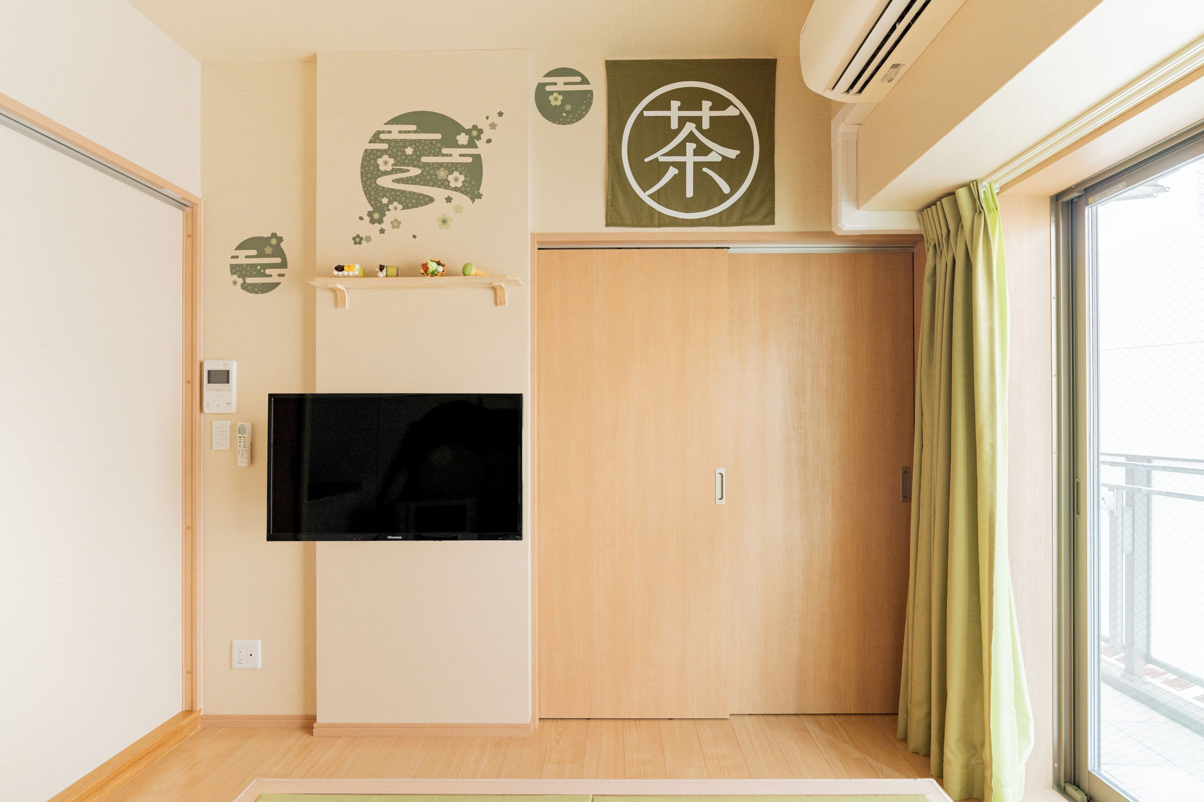 Een kamer met een aan de muur bevestigde televisie, een houten garderobe, een plank met decoratieve objecten en een raam met gordijnen.