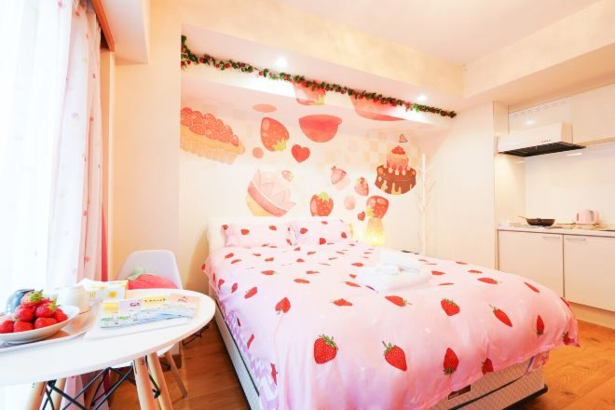 Ein Schlafzimmer mit einem Bett, das mit einer decke mit Erdbeermuster bezogen ist, einem kleinen Tisch mit einer Schale Erdbeeren und einer Wand mit Obstbildern.