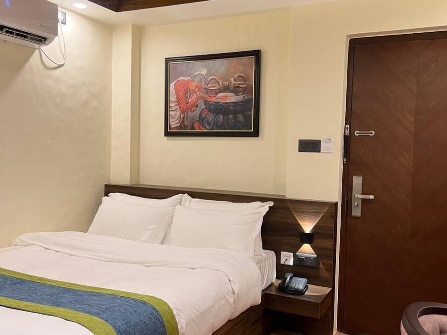 A hotel room with a bed, a nightstand, a wall-mounted air conditioner, a door, and a framed artwork on the wall.