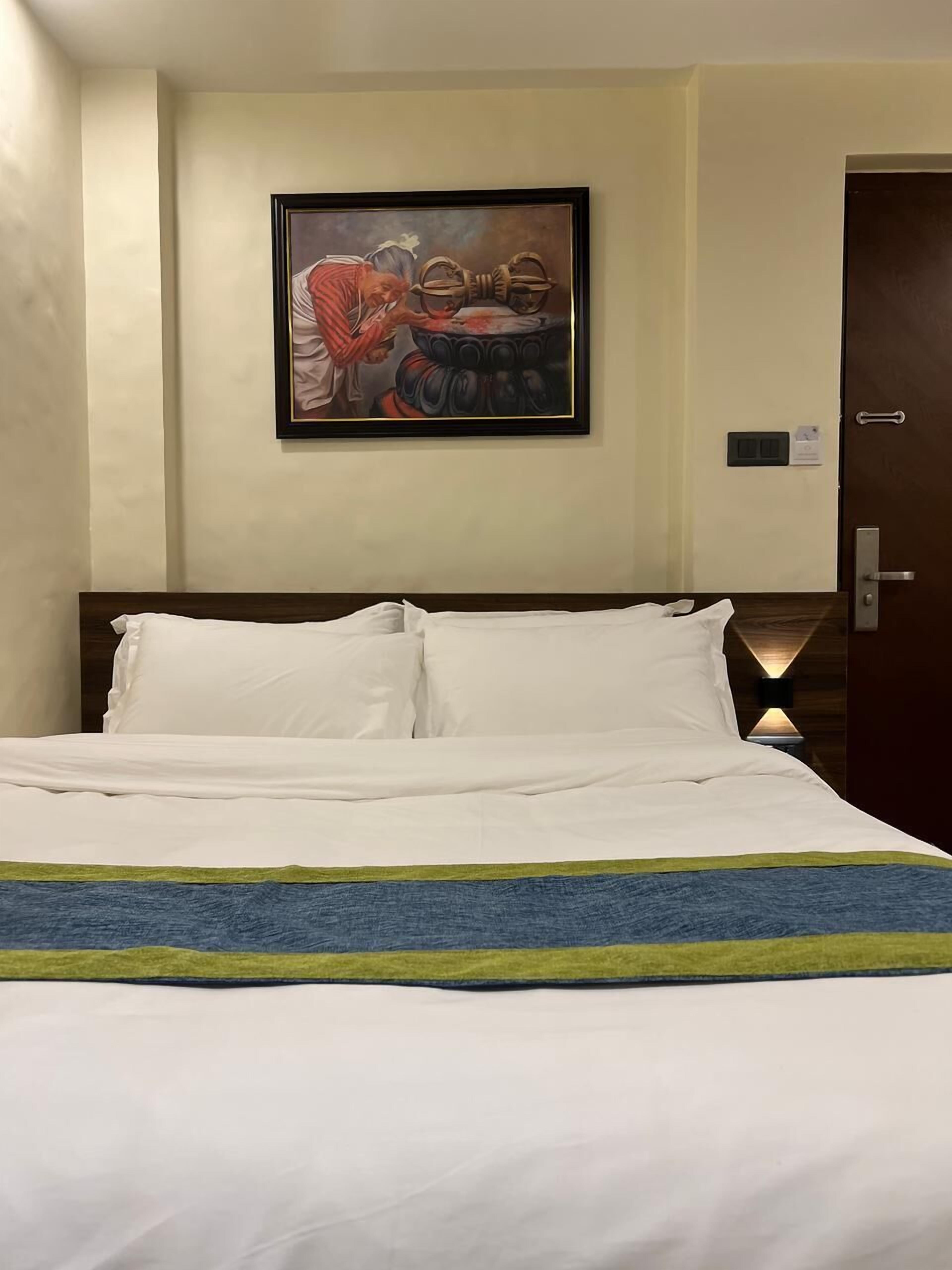 Ein Hotelzimmer mit Bett, einem gerahmten Gemälde an der Wand, einer Tür mit Griff und einem Lichtschalter.