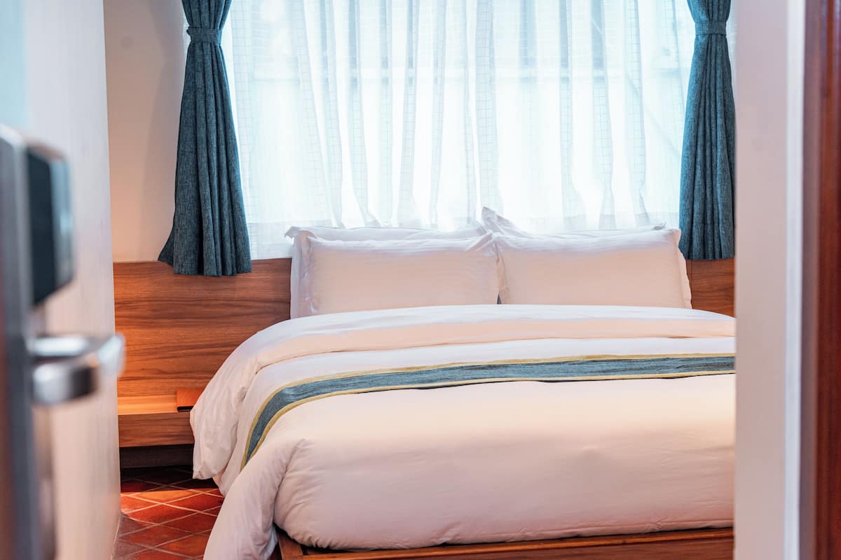 Un lit bien fait, avec du linge de lit blanc orné d’une bande bleue, une tête de lit en bois et une fenêtre agrémentée de rideaux légers.