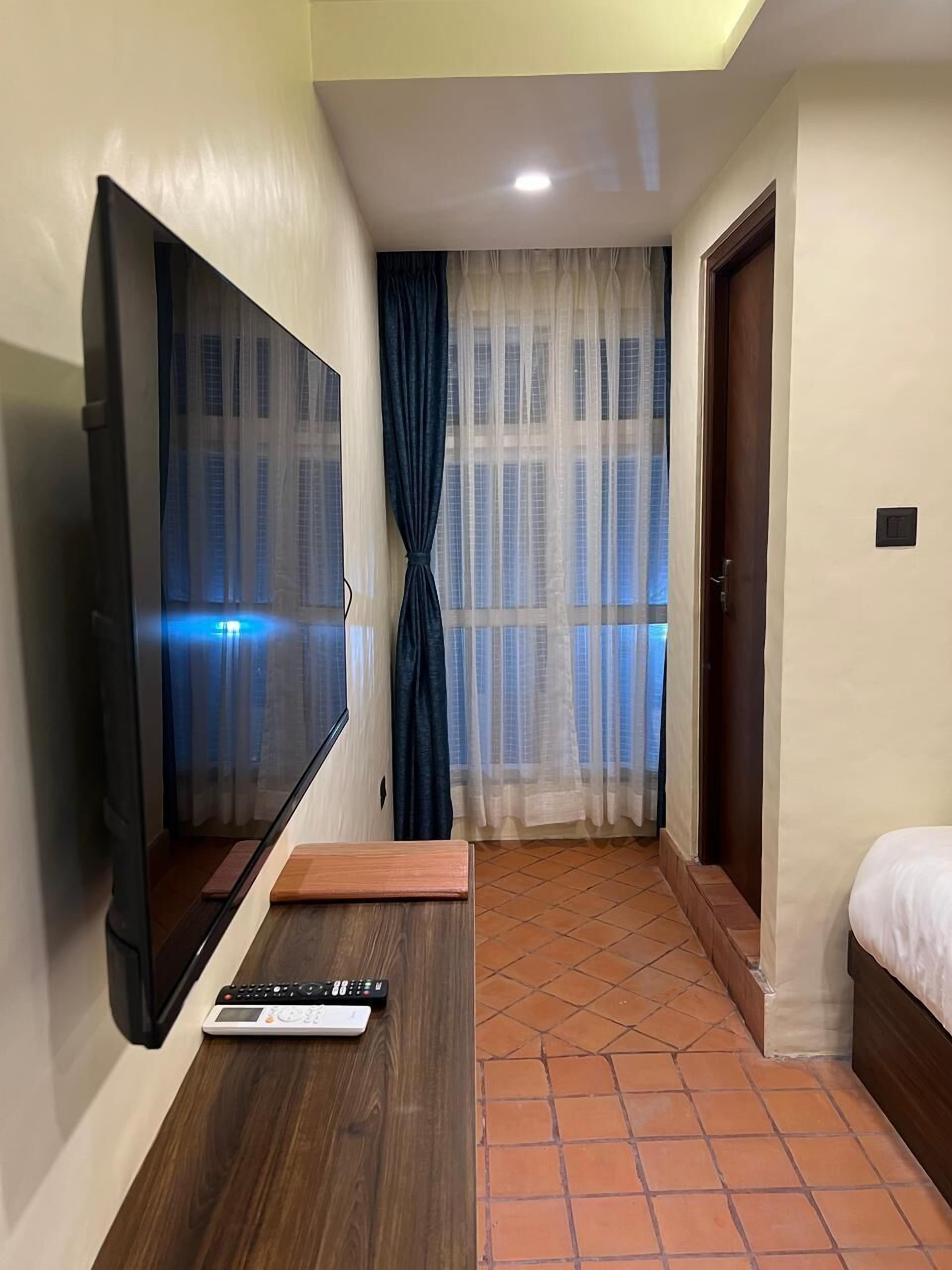 A hotel room with a TV mounted on the wall, a bed, a small wooden table, and a window with curtains.
