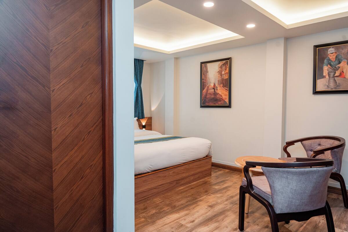 Une chambre d’hôtel moderne avec un plancher en bois, un lit agrémenté d’une touche de bleu, une petite table ronde, deux chaises et deux œuvres d’art encadrées accrochées aux murs.