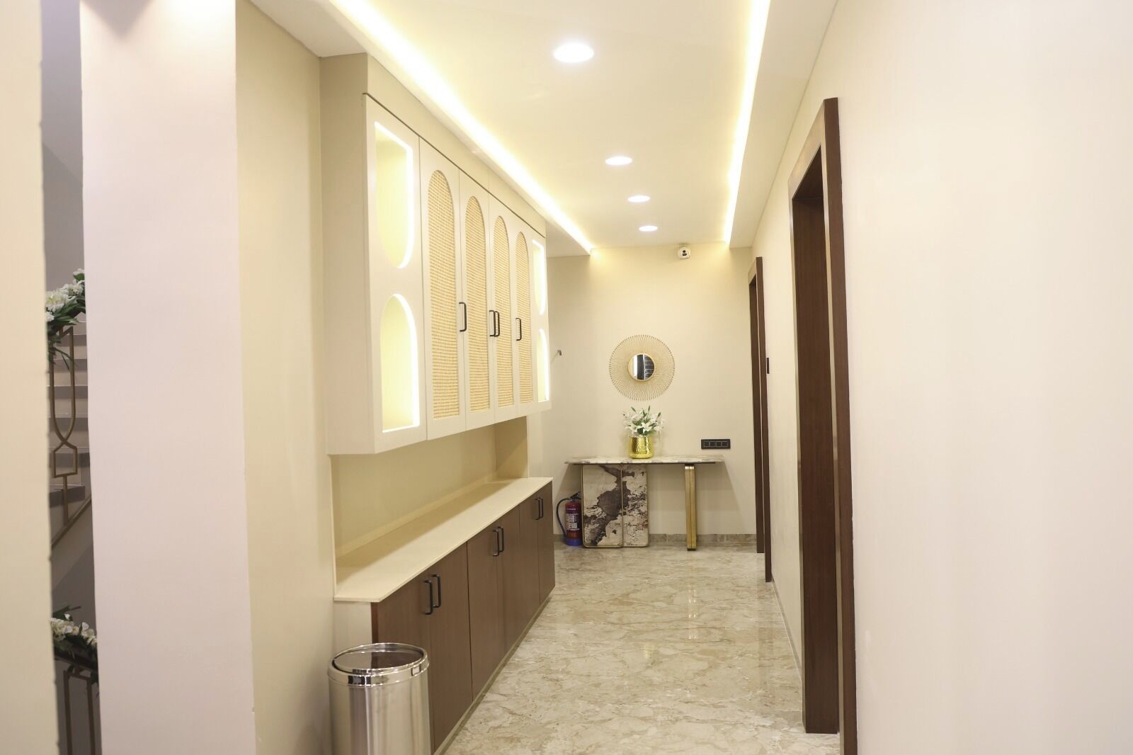 Un pasillo con piso de mármol, un mueble de pared con paneles iluminados, una pequeña mesa con un florero y un cesto de basura.