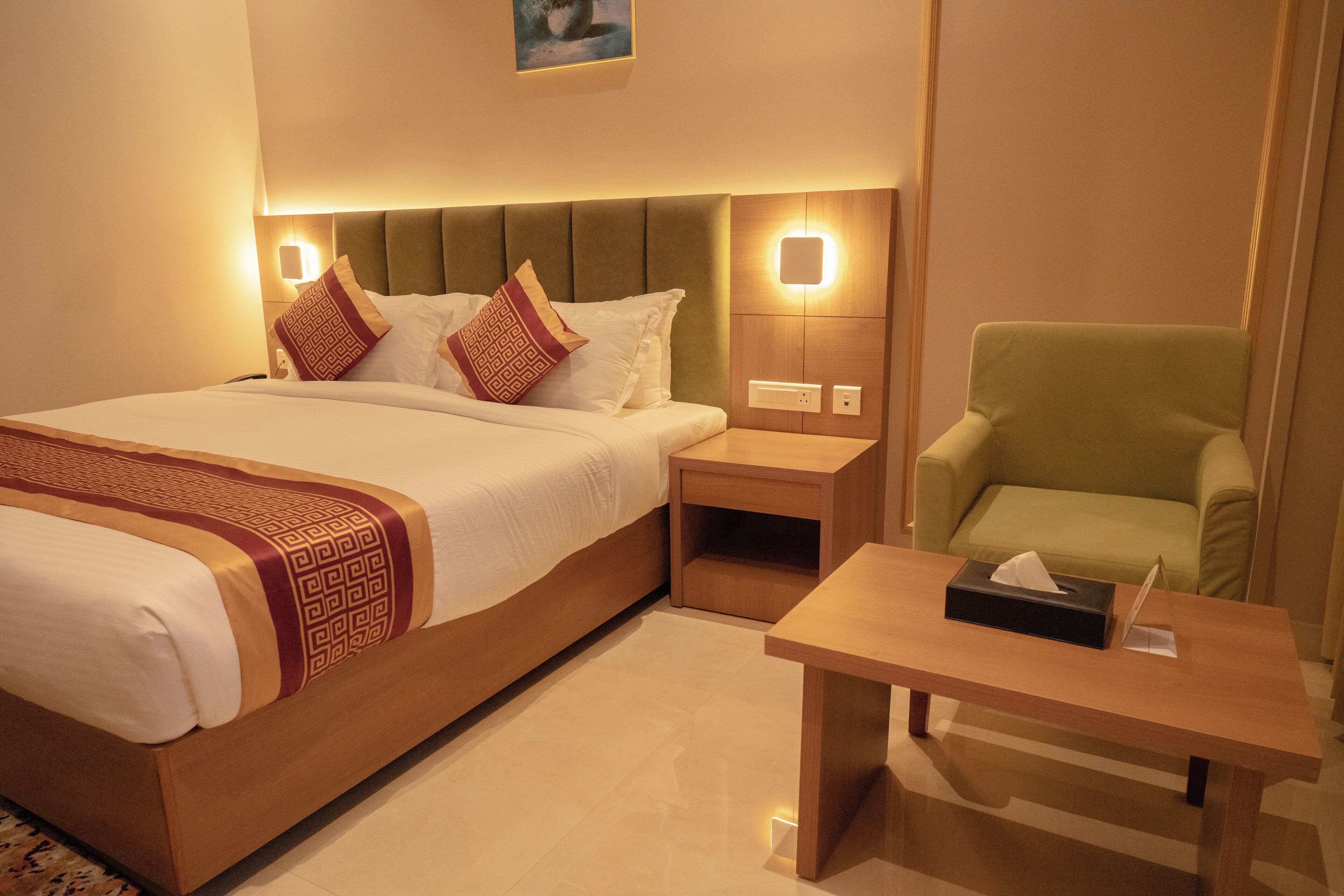 Ein Hotelzimmer mit einem großen Bett, einem grünen Sessel, einem Nachttisch aus Holz und einem Beistelltisch mit Taschentuchbox.