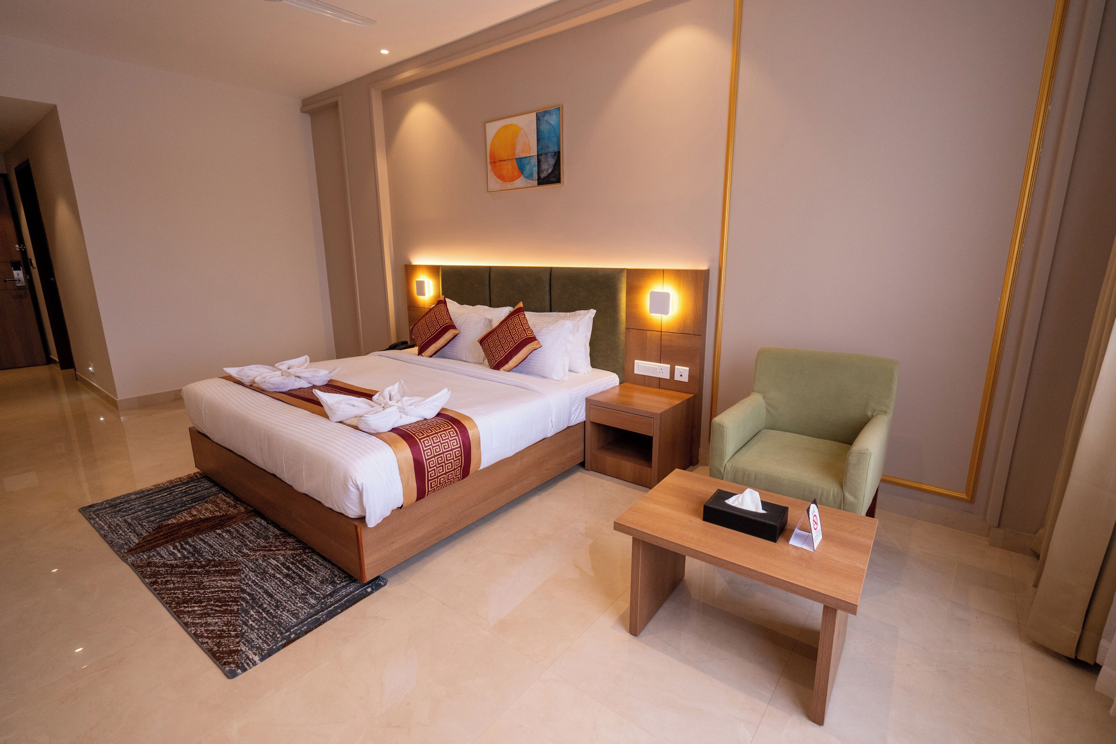 A modern hotel room with a large bed, bedside tables, a green armchair, and a small table with a tissue box.