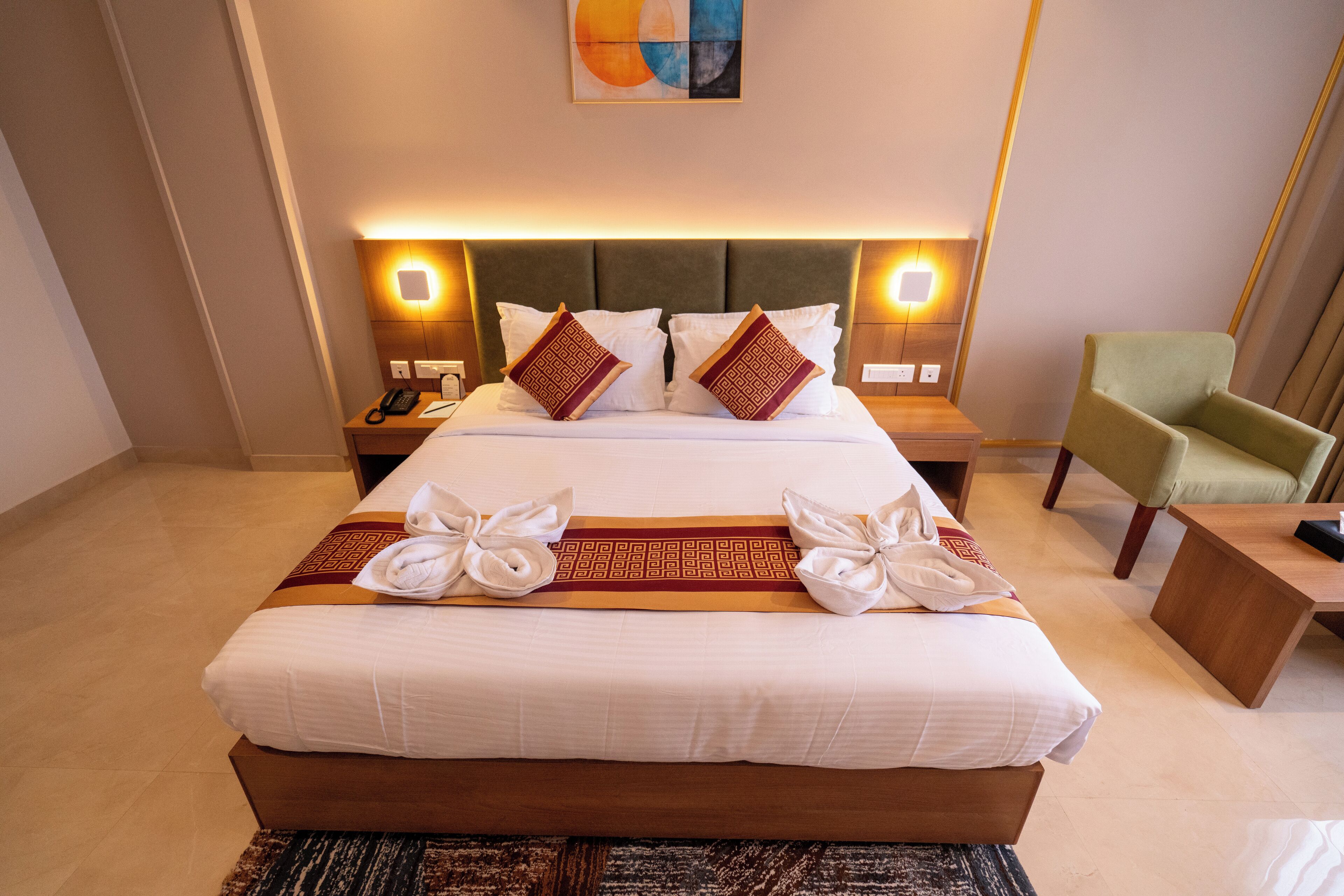 A neatly made bed with white linens and decorative pillows, a bedside table with a lamp, and a green armchair in a modern hotel room.