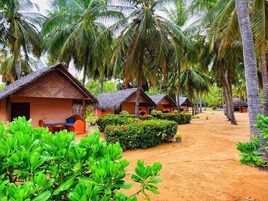 Fileira de cabanas com telhados de palha, separadas por um caminho de areia, rodeadas por vegetação exuberante e palmeiras.