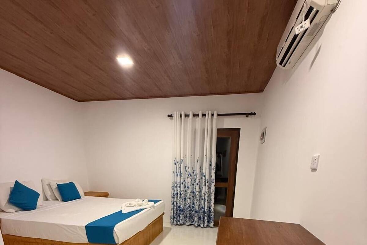 A bedroom with a wooden bed, a bedside table, a wooden bench, a large window with curtains, and a ceiling fan.