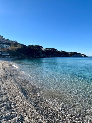 Ein Kiesstrand mit klarem Wasser, ein Restaurant namens "La Roca & Restaurant" auf der linken Seite und Gebäude, die sich an einem Hügel erheben.