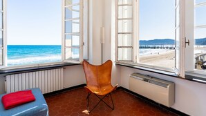 Une pièce dotée de grandes fenêtres donnant sur une plage, d’un fauteuil et d’un radiateur.