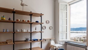 Une pièce comprenant une table à manger en bois, une étagère haute ornée de divers objets décoratifs, une chaise blanche et une vue sur une plage, visible à travers une grande fenêtre.