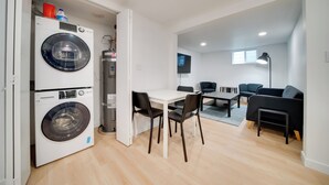 Un espace de vie compact comprenant un lave-linge, une table à manger avec des chaises, un canapé et un téléviseur fixé au mur.