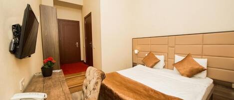 Ein Hotelzimmer mit Bett, Schreibtisch, Telefon und Fernbedienung, einem an der Wand befestigten Fernseher und einer Tür zu einem anderen Raum.