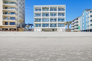 A multi-story hotel with balconies, located on a sandy beach.