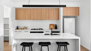 A modern kitchen with white cabinetry, a central island with bar stools, and a built-in oven. - St Helens I Stunning Family Townhouse in Geelong (Rippleside)