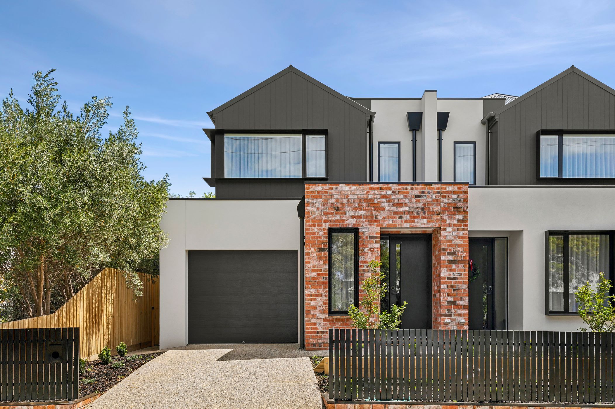 Une maison moderne à deux étages, dotée d’un mur d’accent en briques, de grandes fenêtres et d’une porte de garage de couleur sombre.