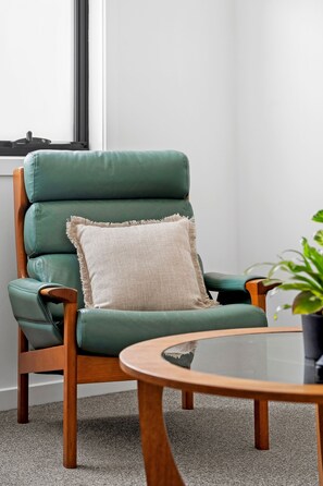 Een groene leren fauteuil met een beige kussen, geplaatst naast een ronde glazen tafel met houten poten, tegen een witte muur met een raam.